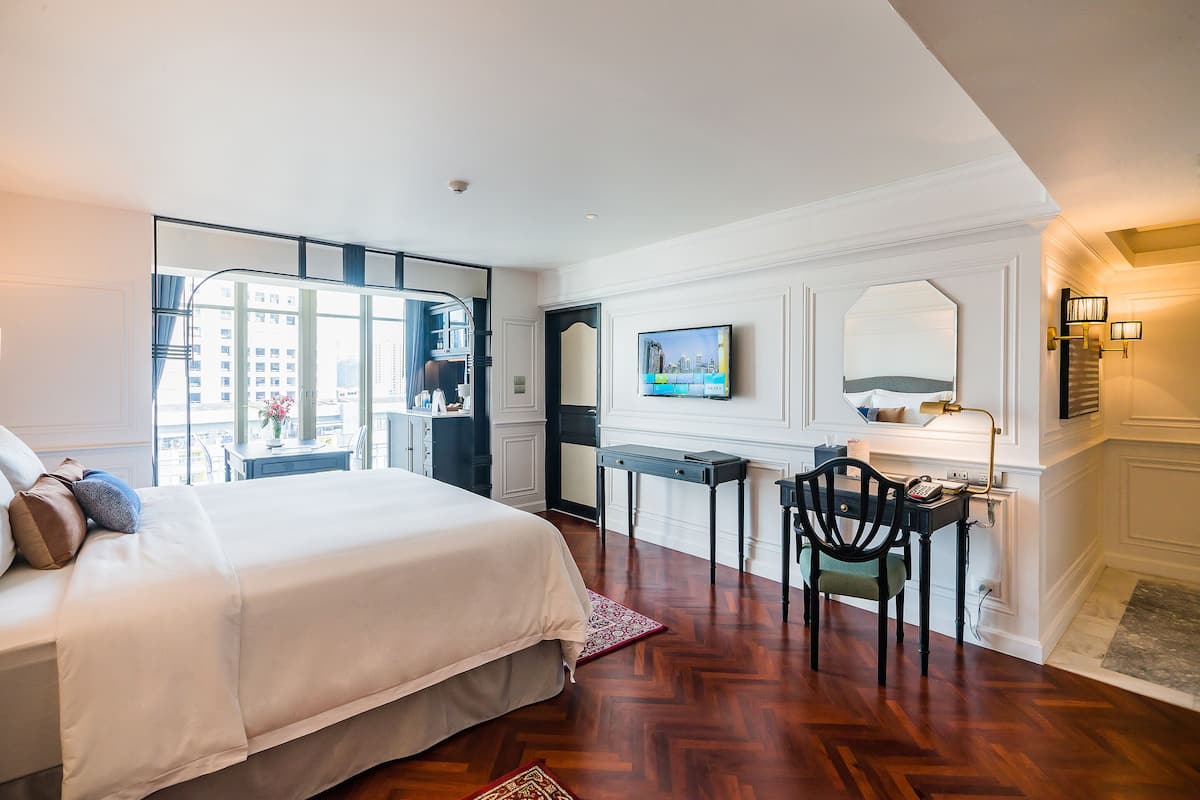 A hotel room with a bed, a desk, a chair, a TV, and a view of the city through large windows.