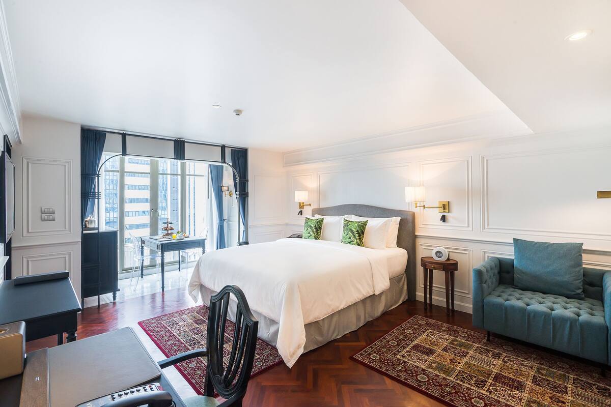 A hotel room with a large bed, a sitting area with a sofa and armchair, a desk with a laptop, and a view of the city through the window.