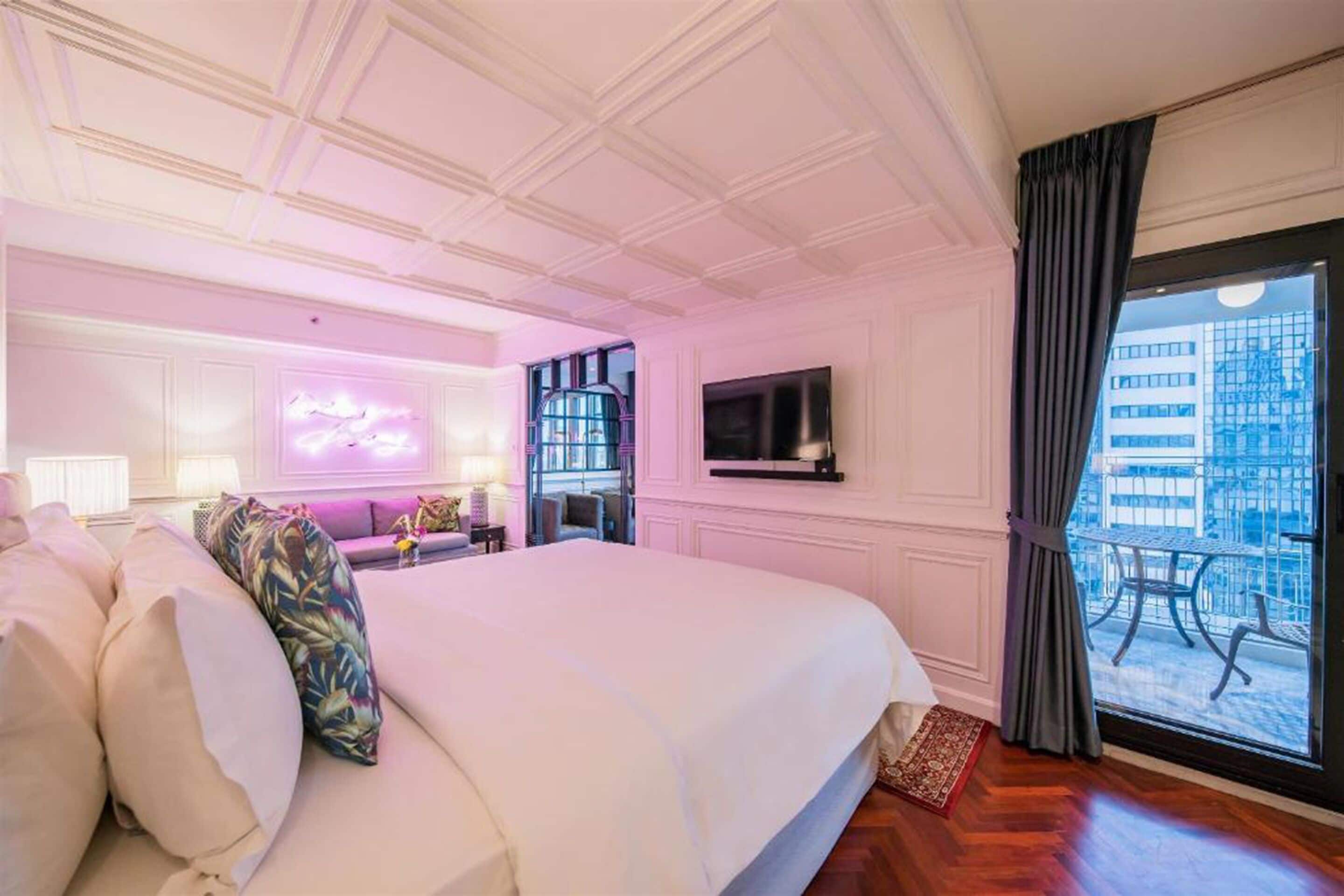 Ein Hotelzimmer mit einem großen Bett, einem Fernseher, einer Neonreklame und einem Balkon mit Gartenmöbeln.