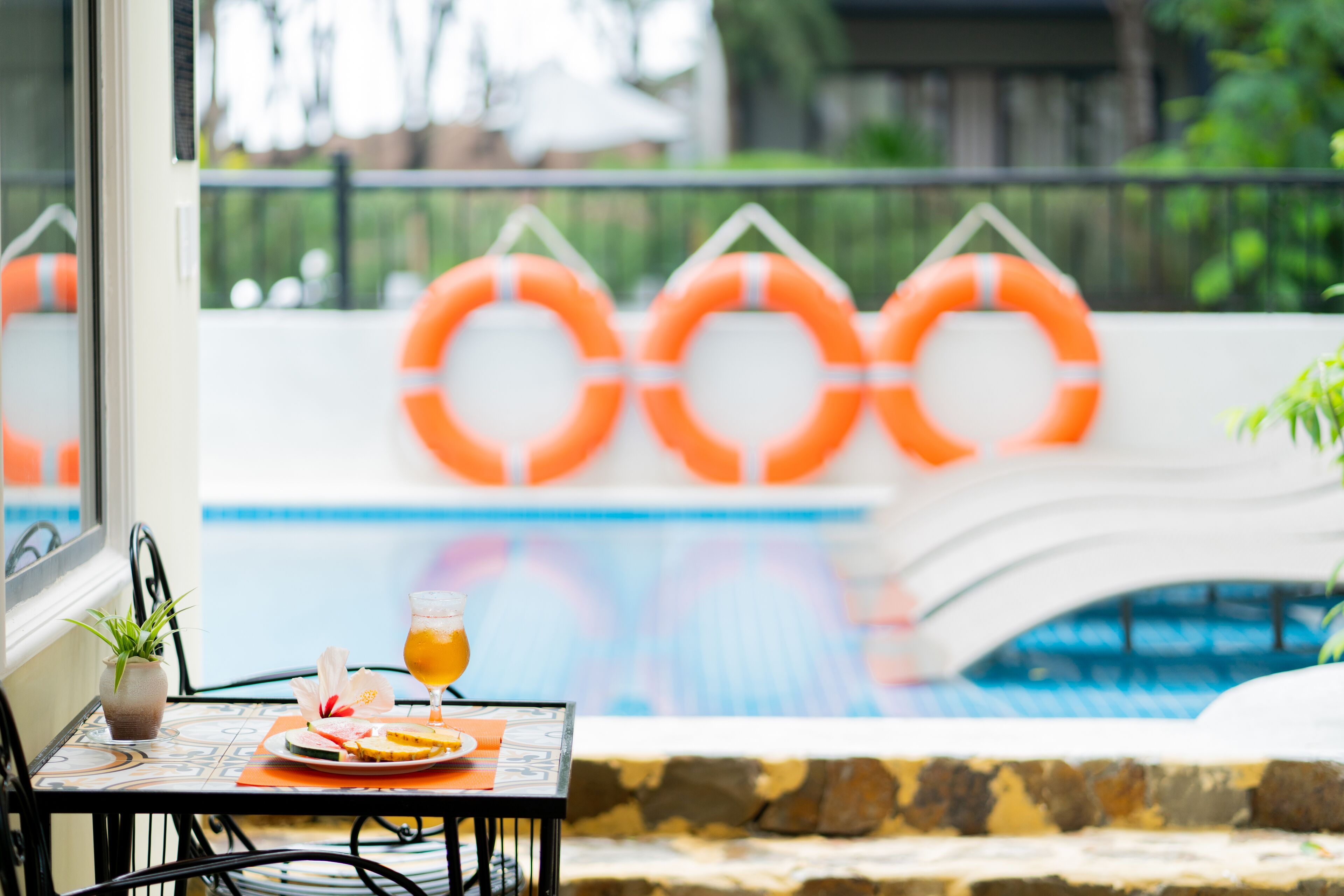 A poolside table with a drink and fruit, a potted plant, and orange lifebuoys in the background.