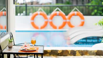 A poolside table with a drink and fruit, a potted plant, and orange lifebuoys in the background.