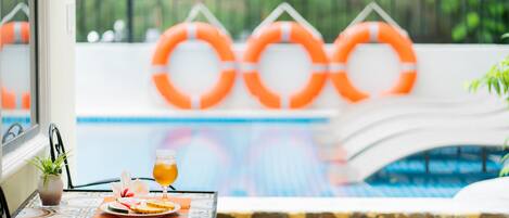 A poolside table with a drink and fruit, a potted plant, and orange lifebuoys in the background.