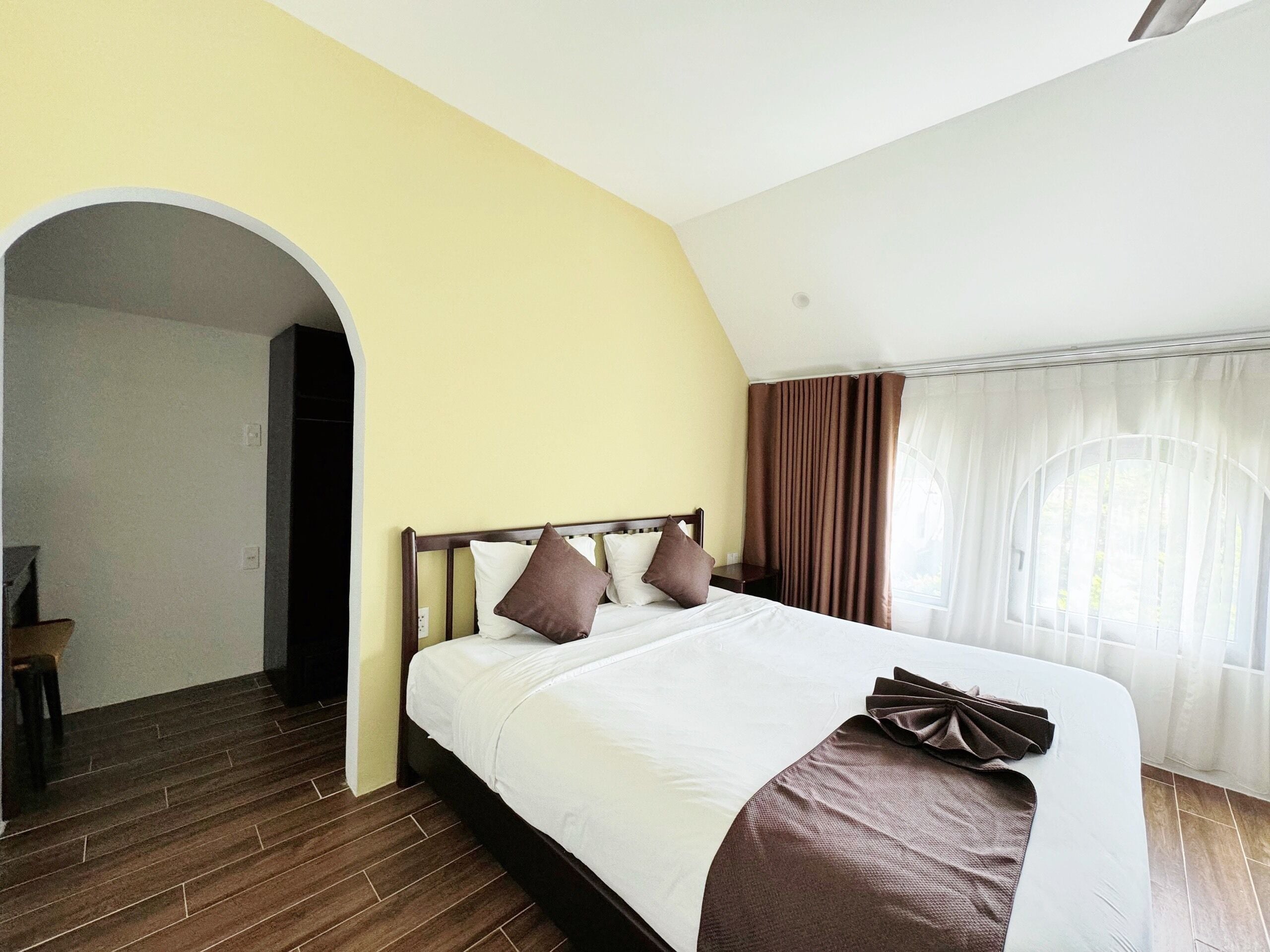A hotel room with a large bed, a wooden floor, a yellow wall, and a window with curtains.