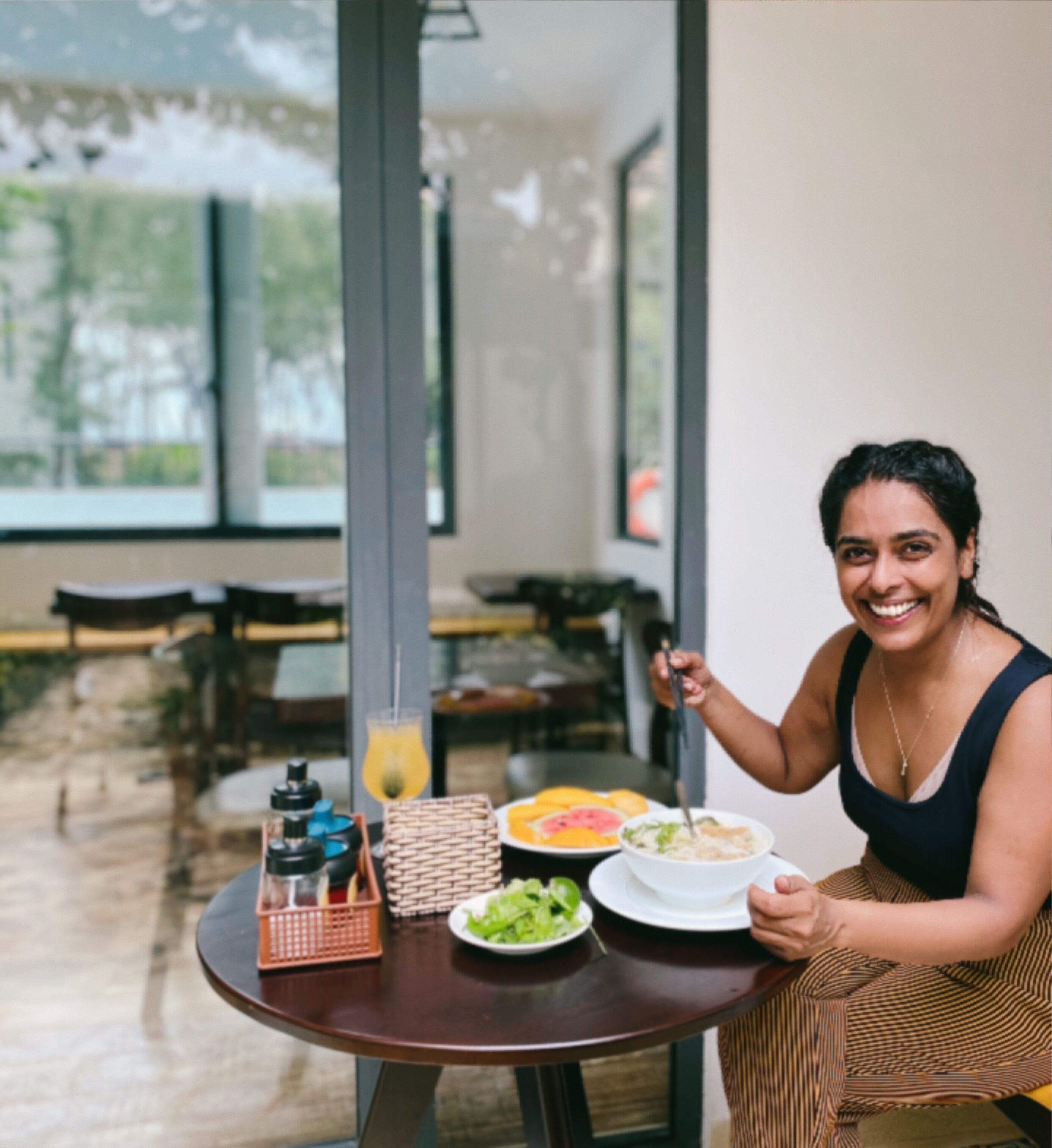 Eine Person sitzt an einem runden Tisch mit Mahlzeit, Getränk und Beilagen. Im Hintergrund sind große Fenster und ein Blick auf Grünflächen zu sehen.