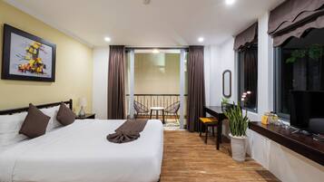 A hotel room with a bed, a desk, a chair, a potted plant, and a balcony with curtains.