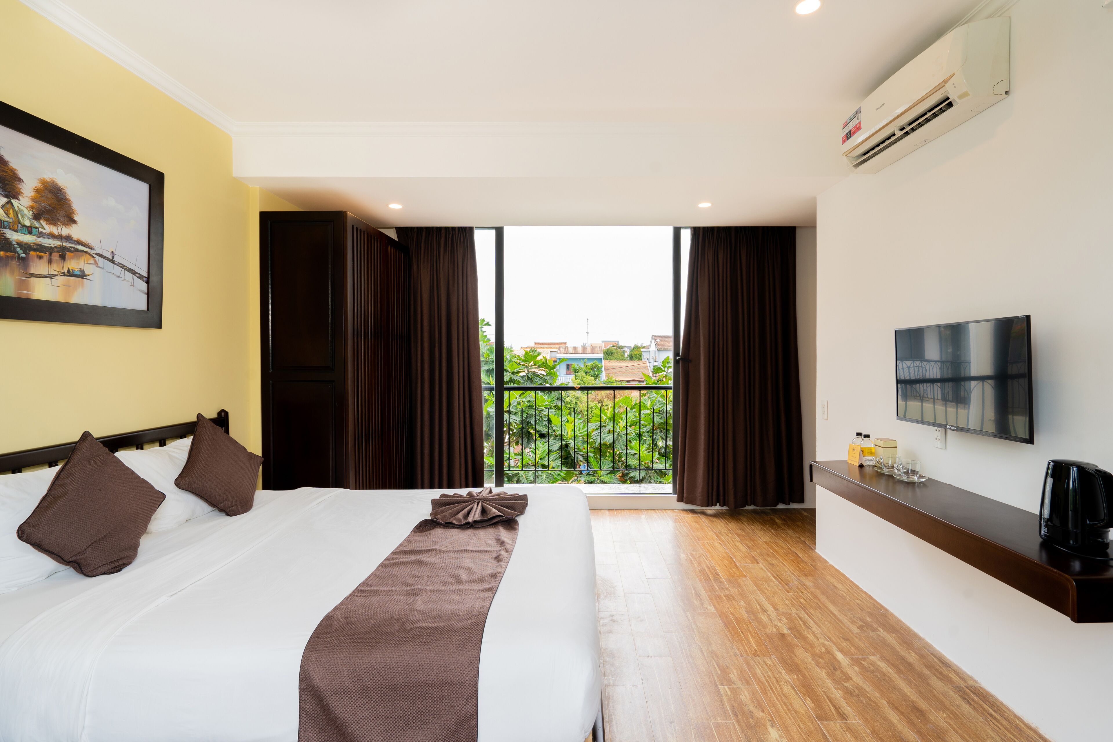 Habitación de hotel con cama, televisión, cafetera y balcón con vistas a zonas verdes y edificios.
