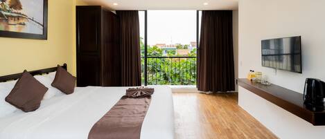 Habitación de hotel con cama, televisión, cafetera y balcón con vistas a zonas verdes y edificios.