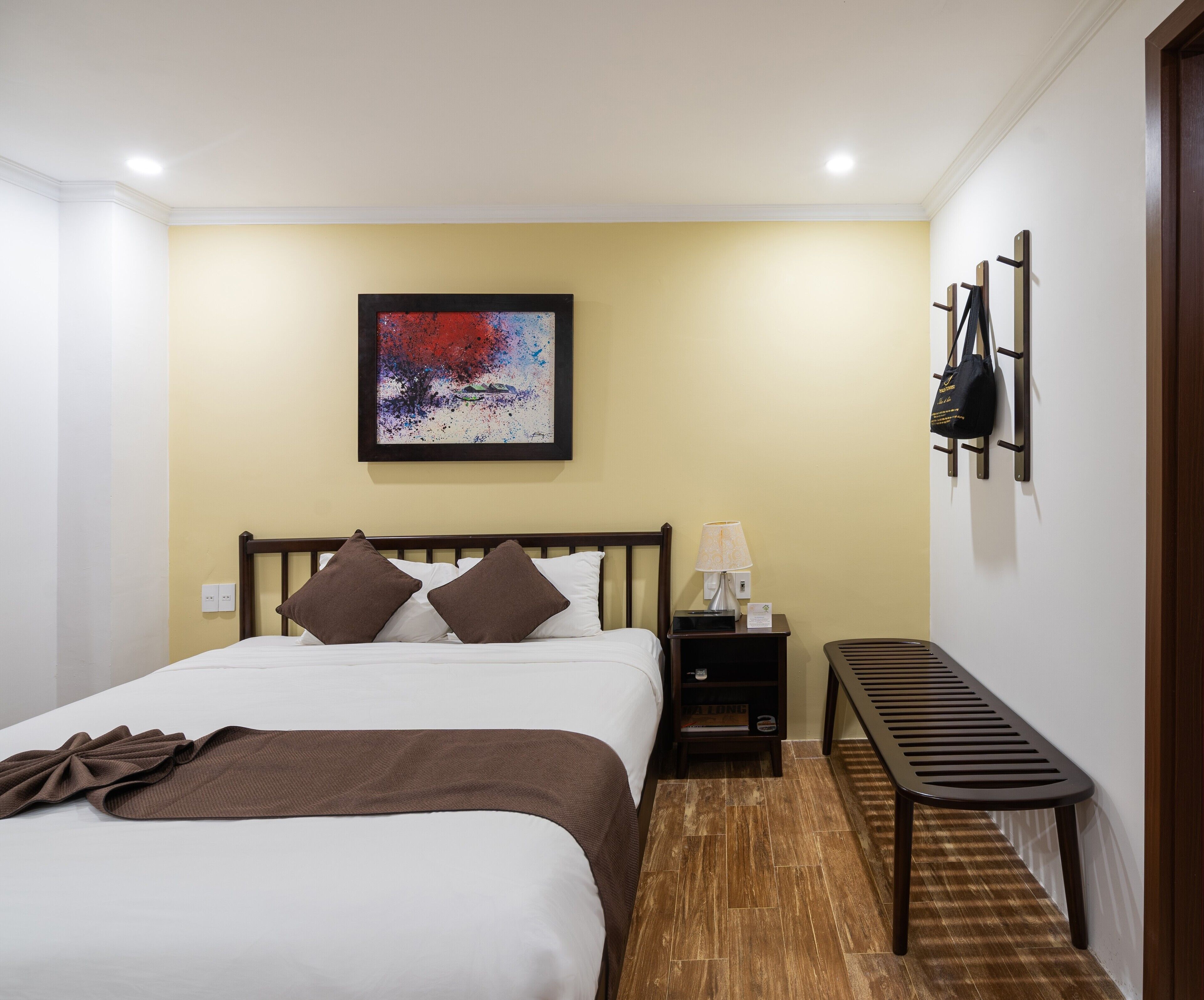 A hotel room with a bed, a nightstand, a lamp, a bench, and a wall-mounted coat rack.