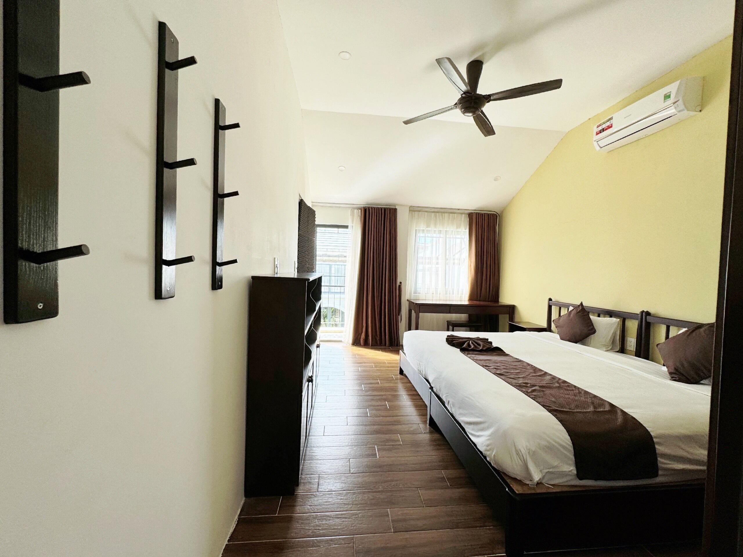 Habitación de hotel con cama, escritorio, ventilador de techo y ventana con cortinas.