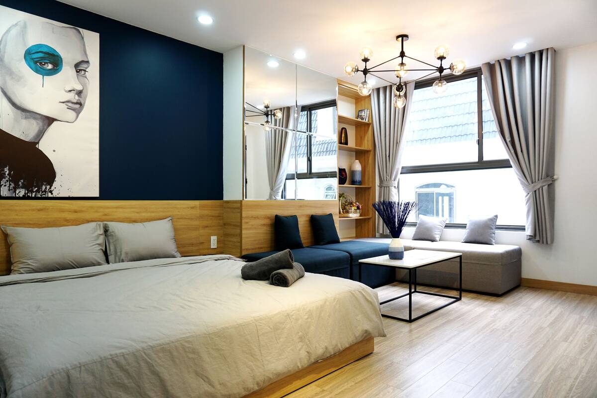 A modern hotel room with a large bed, a sofa, a small table, and a large window with curtains.