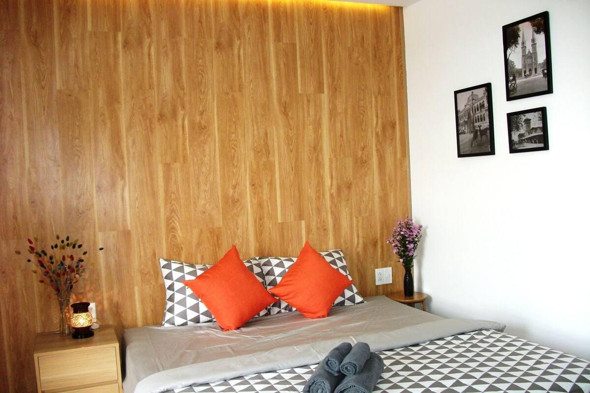 A neatly arranged hotel room with a wooden headboard, a bed with patterned bedding, two orange pillows, a nightstand with a lamp, and a vase with flowers.