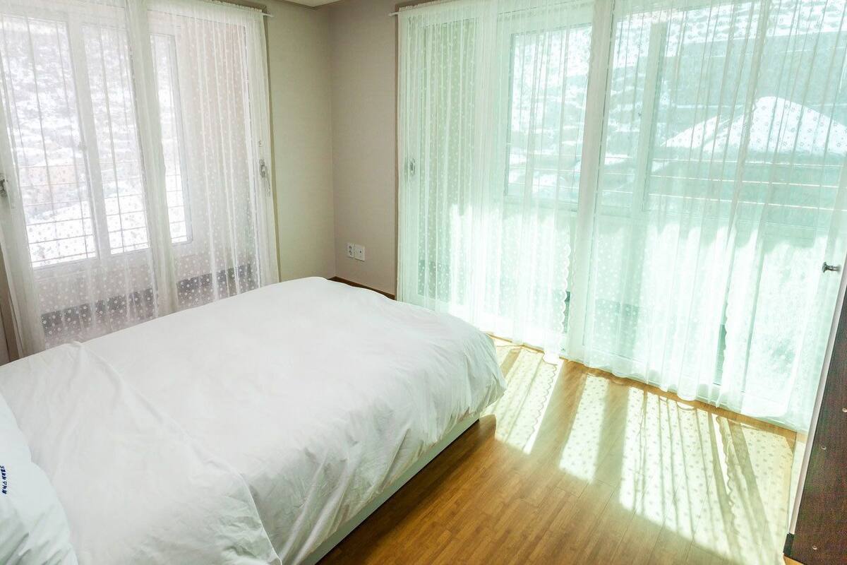 A bedroom with a bed, two windows with sheer curtains, and a wooden floor.