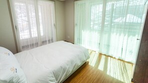 A bedroom with a bed, two windows with sheer curtains, and a wooden floor.