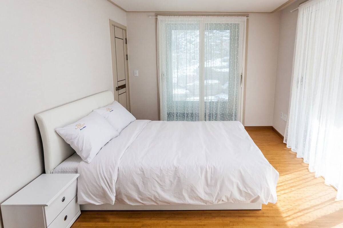 A bedroom with a bed, a nightstand, a wardrobe, and a large window with sheer curtains.