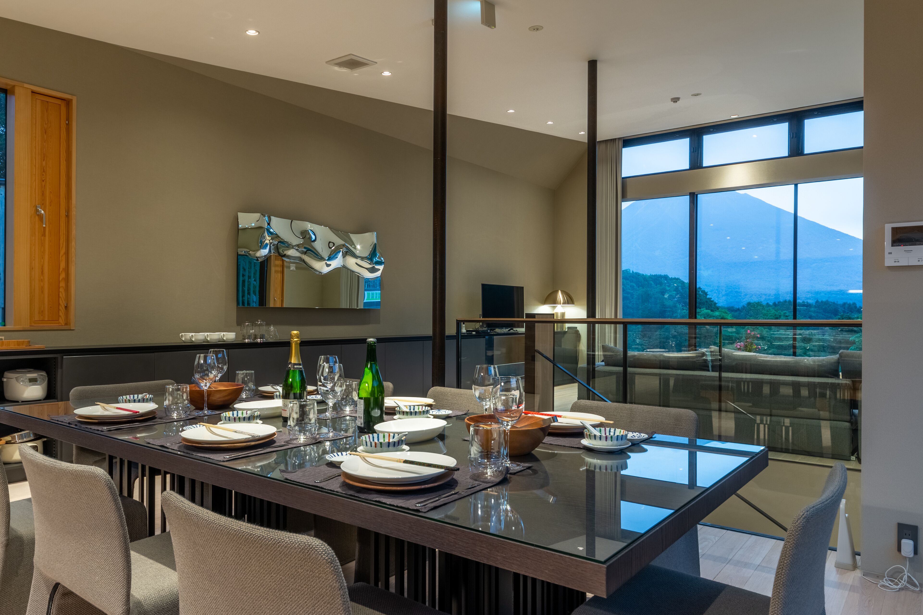 A modern dining area with a glass table, chairs, and a view of a mountain through large windows.