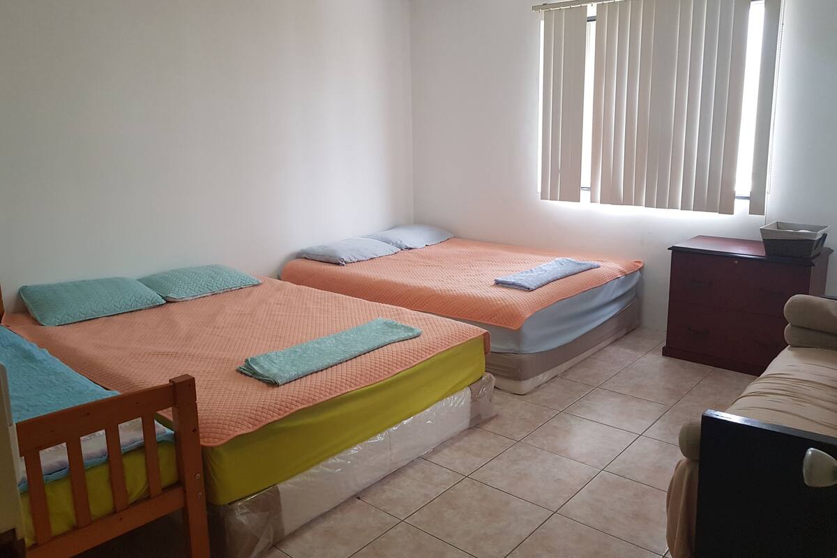 Ein Zimmer mit zwei Betten, einem Holzstuhl, einem kleinen Tisch und einem Fenster mit Jalousien.