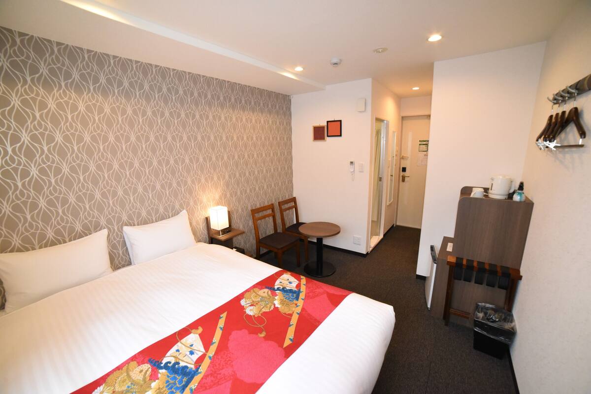 Una habitación de hotel con una cama grande, un escritorio, una silla, una mesita, un perchero y un cubo de basura.