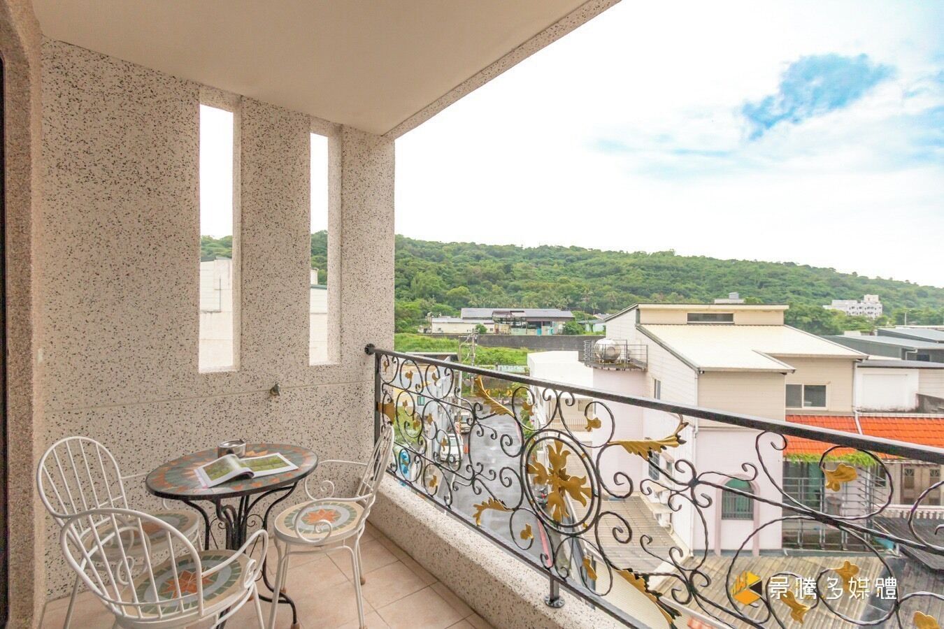 A balcony with a table and chairs, a decorative railing, and a view of buildings and greenery.