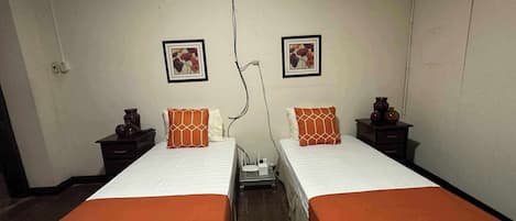 A room with two beds, each with an orange bedspread and white bedding. There are two nightstands with decorative items, and two framed pictures on the wall.