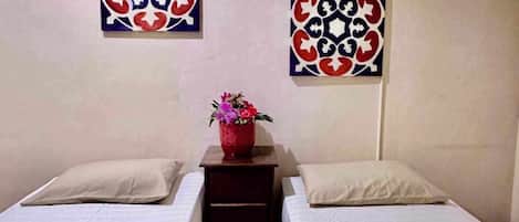 A room with two beds, a small wooden table with a vase of flowers, and two wall-mounted windows.