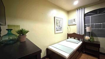 A small, neatly arranged bedroom with a single bed, a bedside table, a nightstand, a potted plant, a framed picture on the wall, and an air conditioning unit.