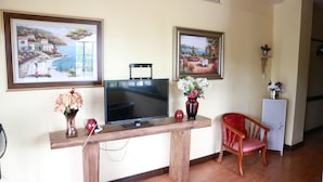Una habitación con un escritorio de madera, un televisor de pantalla plana, dos cuadros enmarcados, una silla roja y un mueble bajo.