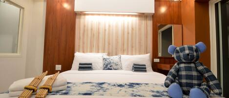 Una habitación de hotel con una cama, almohadas, un osito de peluche y una lámpara de noche.