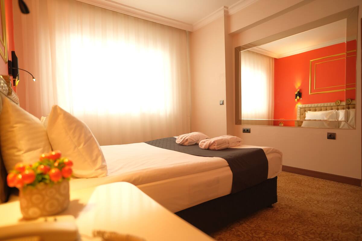 Habitación de hotel con una cama grande, una mesita de noche con un florero y un espejo en la pared, además de una ventana con cortinas.