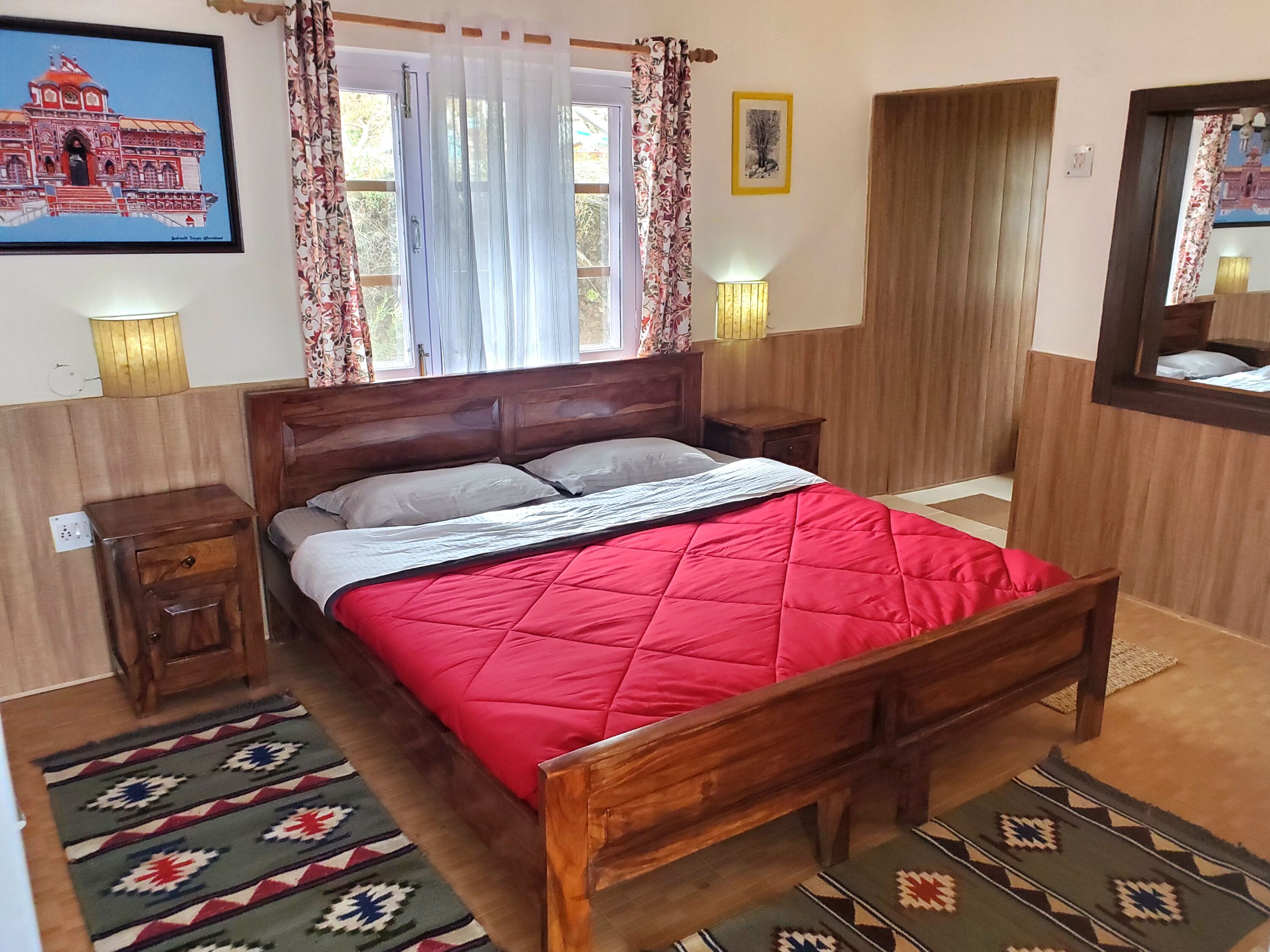 A bedroom with a wooden bed, two bedside tables, a nightstand, a window with curtains, a wall-mounted lamp, and a framed picture.
