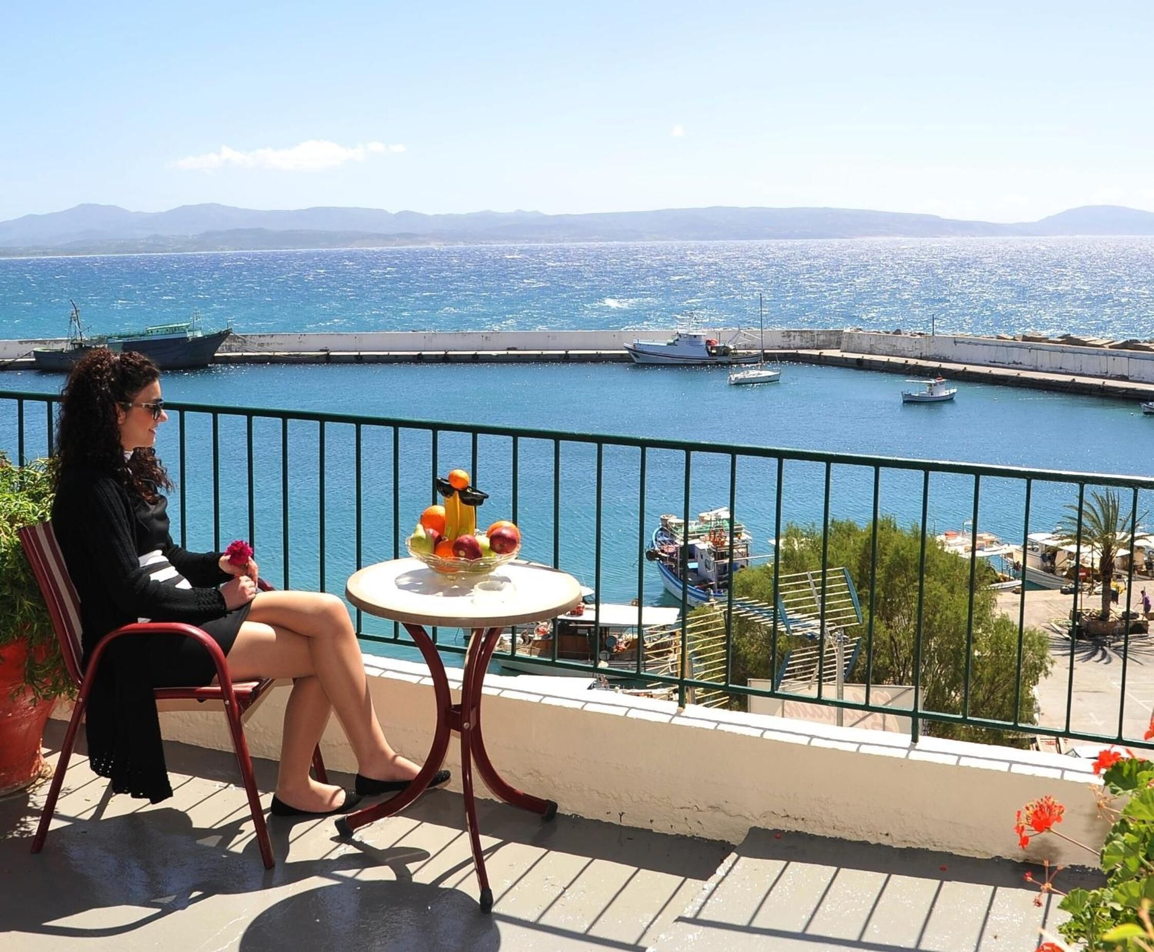 Un balcón con vista a un puerto deportivo, una mesa con fruta y una flor, y una persona sentada.
