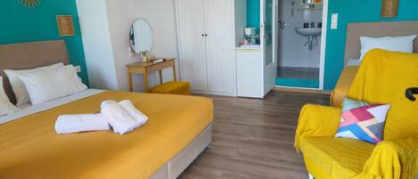 A hotel room with a bed, a yellow armchair, a small table, a wardrobe, and a bathroom visible through an open door.