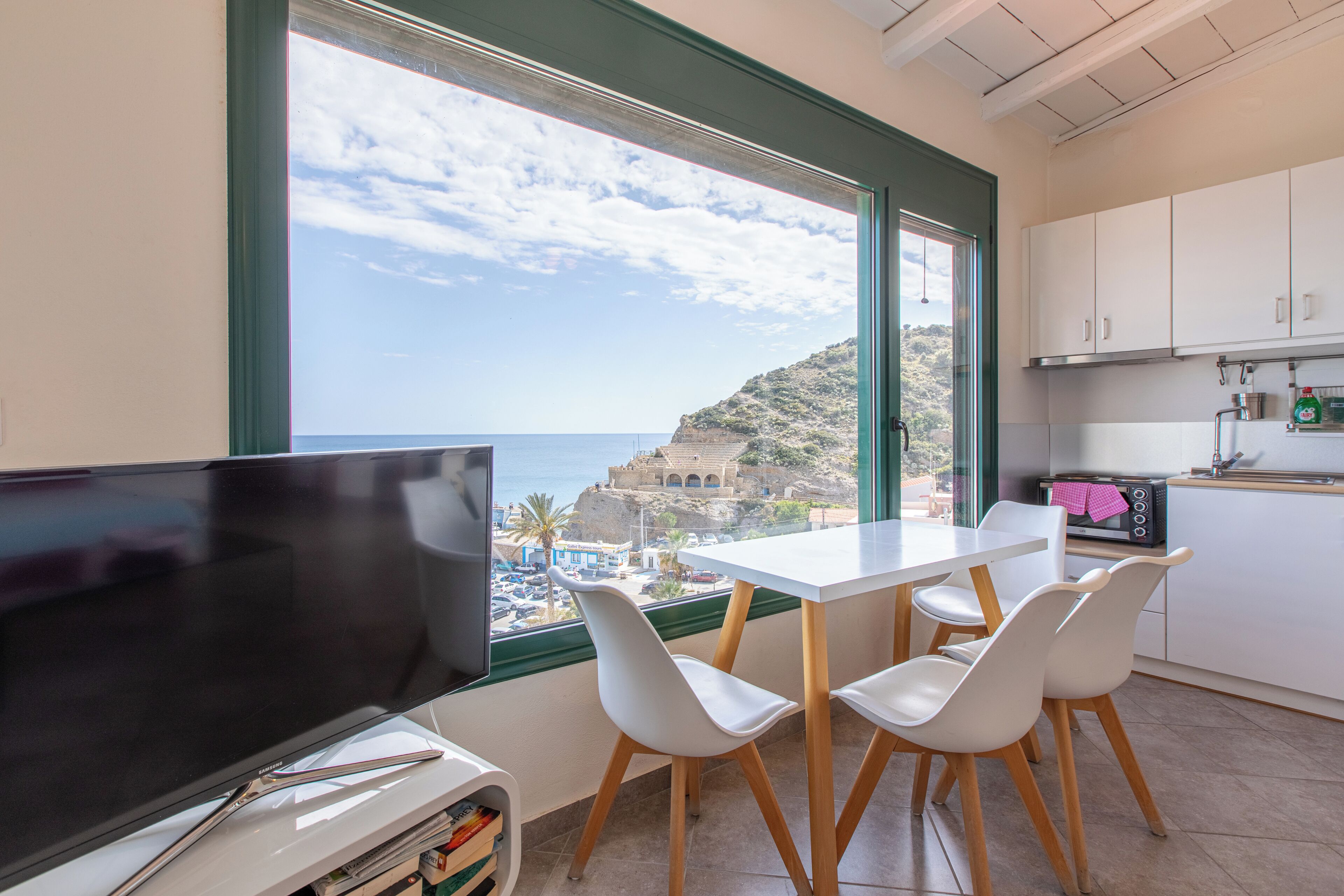 A room with a TV, white chairs, a dining table, a kitchen with white cabinets, and a view of the sea and a cliff.
