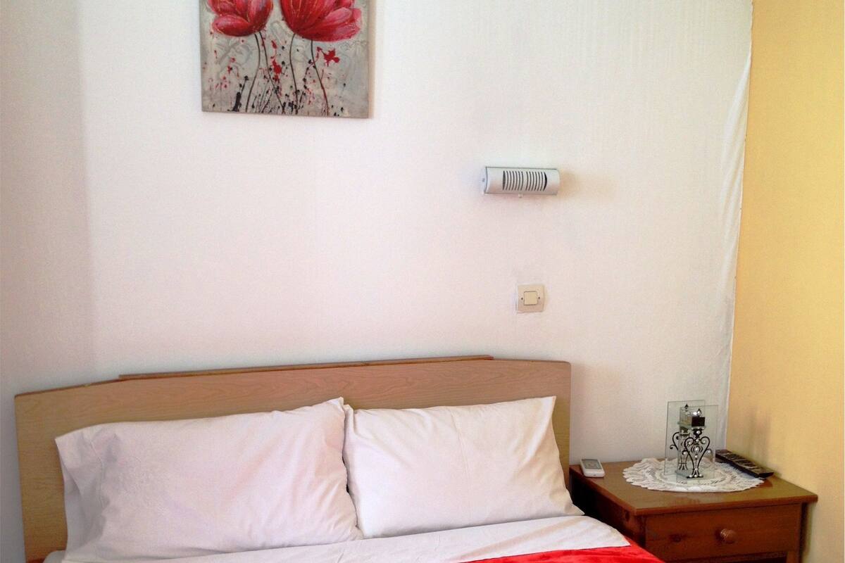 A hotel room with a bed, a nightstand, a painting of flowers, a thermostat, and a bedside table with a lamp and a phone.