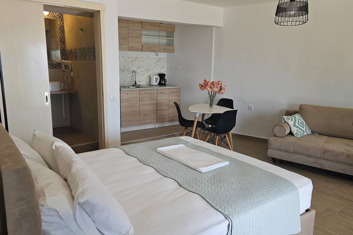 A hotel room with a bed, a sofa, a dining table with chairs, a kitchen area, and a bathroom.