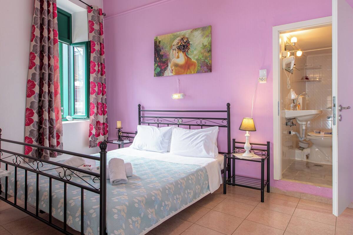 A bedroom with a bed, a nightstand, a lamp, a painting, a bathroom door, and a window with curtains.