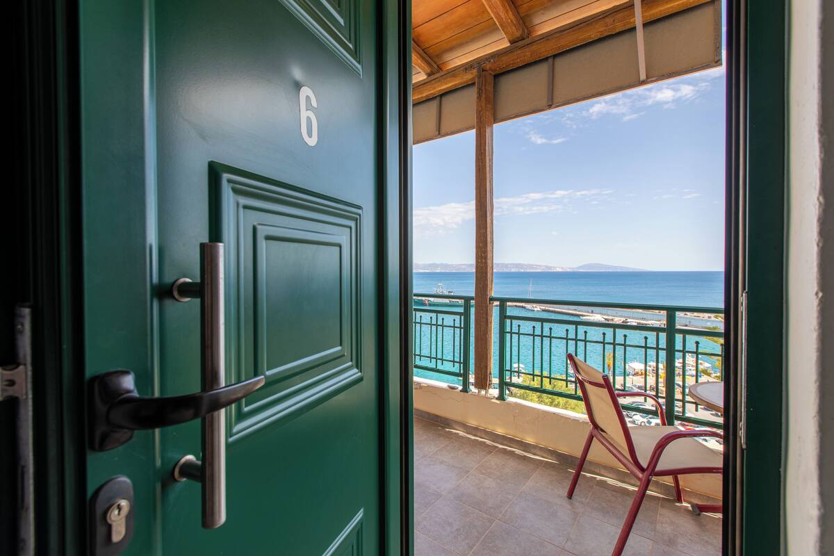 A green door with the number 6, leading to a balcony with a view of the sea and a chair.