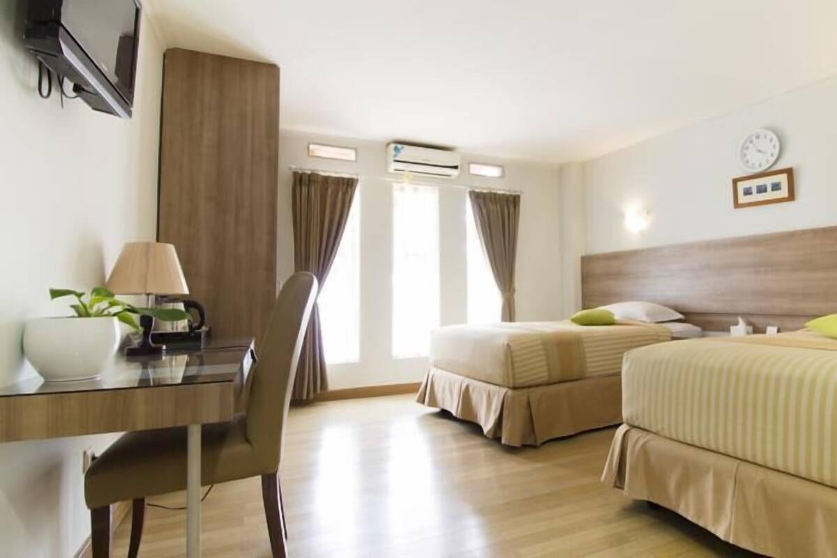 A hotel room with two beds, a desk, a chair, a lamp, a clock, and an air conditioning unit.