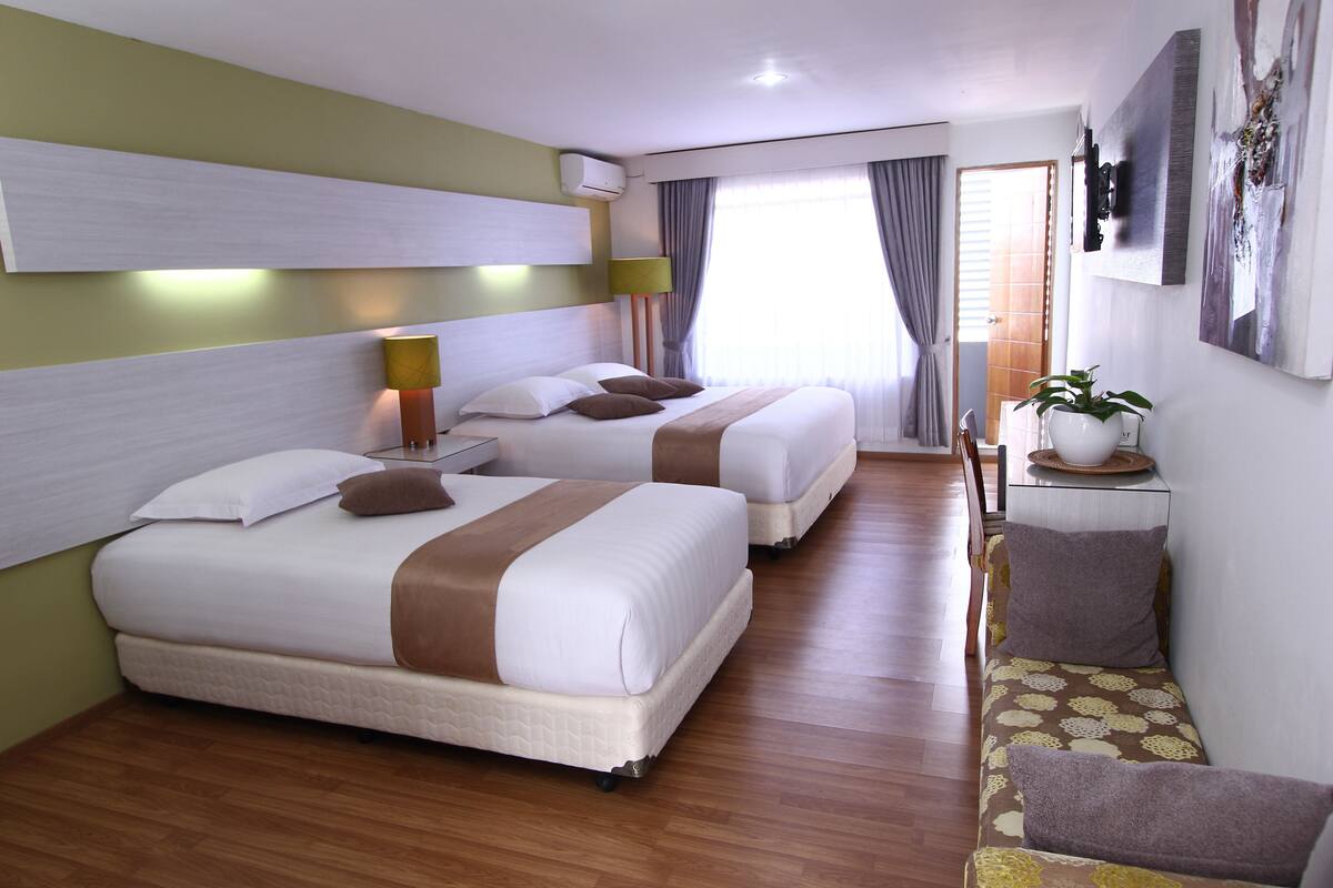 A hotel room with two beds, a sofa, a small table with a plant, and a wall-mounted TV.