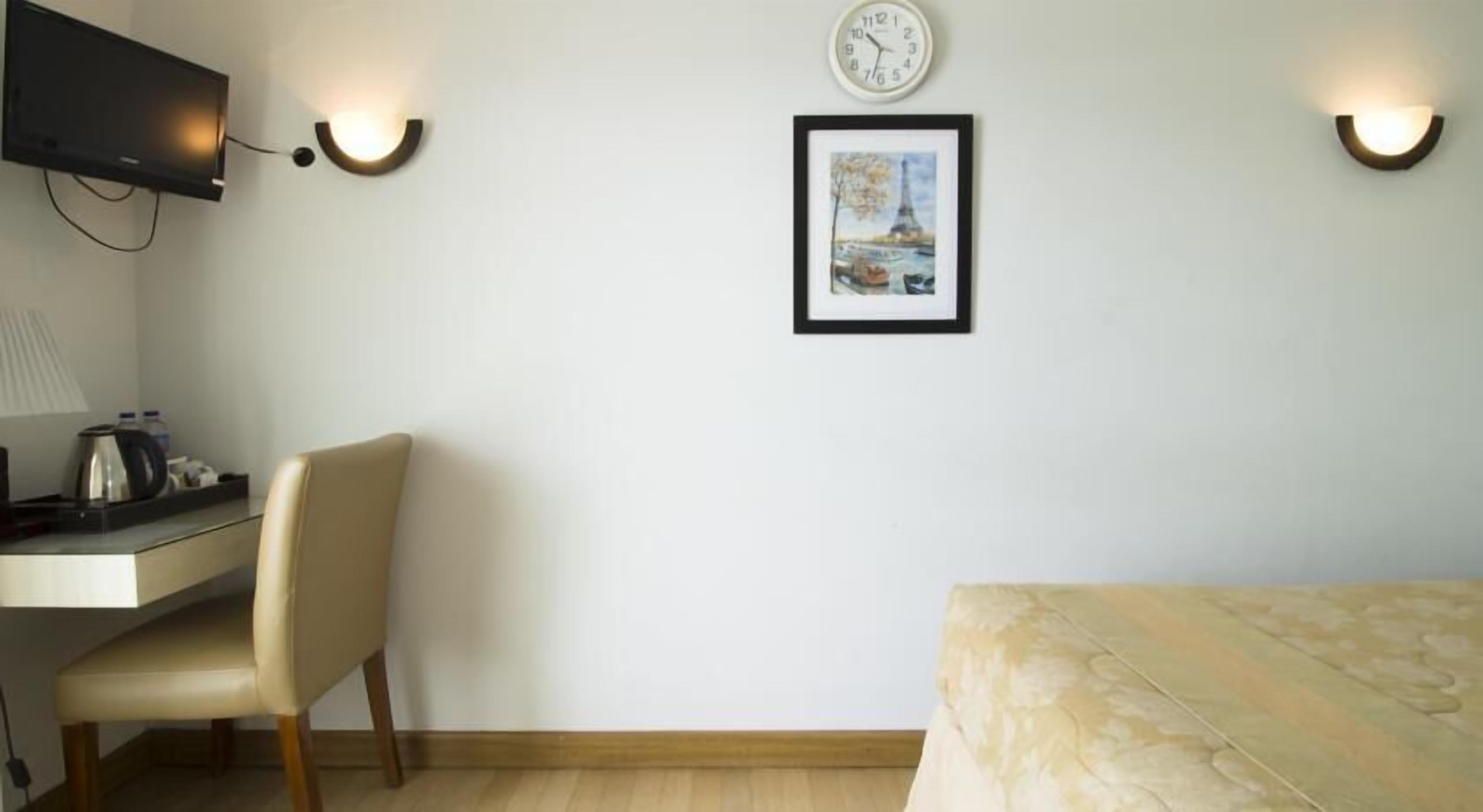 A hotel room with a desk, chair, bed, wall-mounted clock, and a framed picture of the Eiffel Tower.