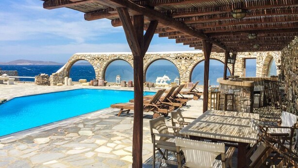 A poolside area with stone flooring, lounge chairs, and a covered seating area with a view of the sea and a stone bridge in the background. - Kanalia Villas (Mykonos)