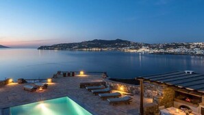 A poolside area with lounge chairs and a dining table, overlooking a calm sea and a cityscape at dusk. - Kanalia Villas (Mykonos)