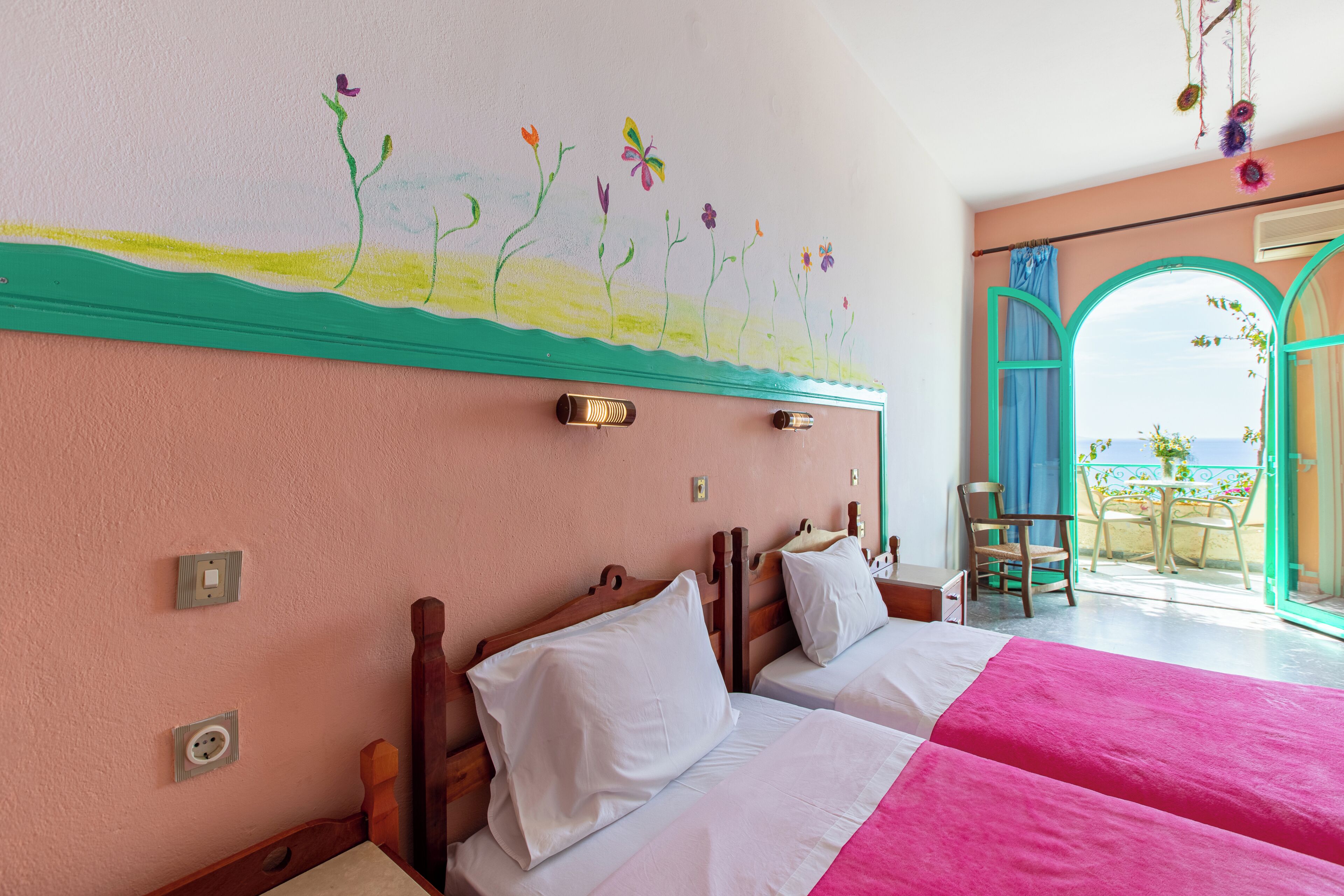 A hotel room with two beds, a painting on the wall, a view through an open door, and a balcony with a table and chairs.