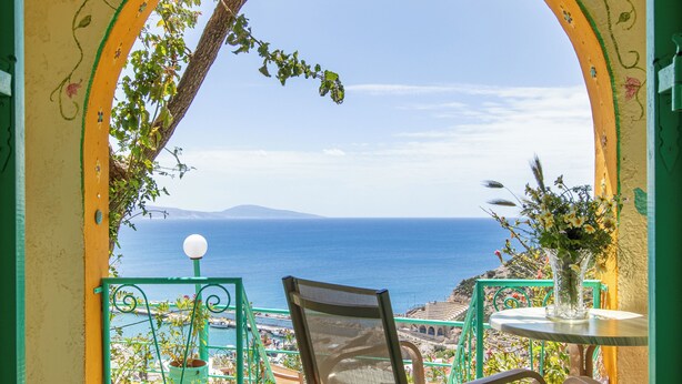 A balcony with a view of the sea, a chair, a table with a vase, and a decorative archway. - Erofili Rooms (Agios Vasileios)