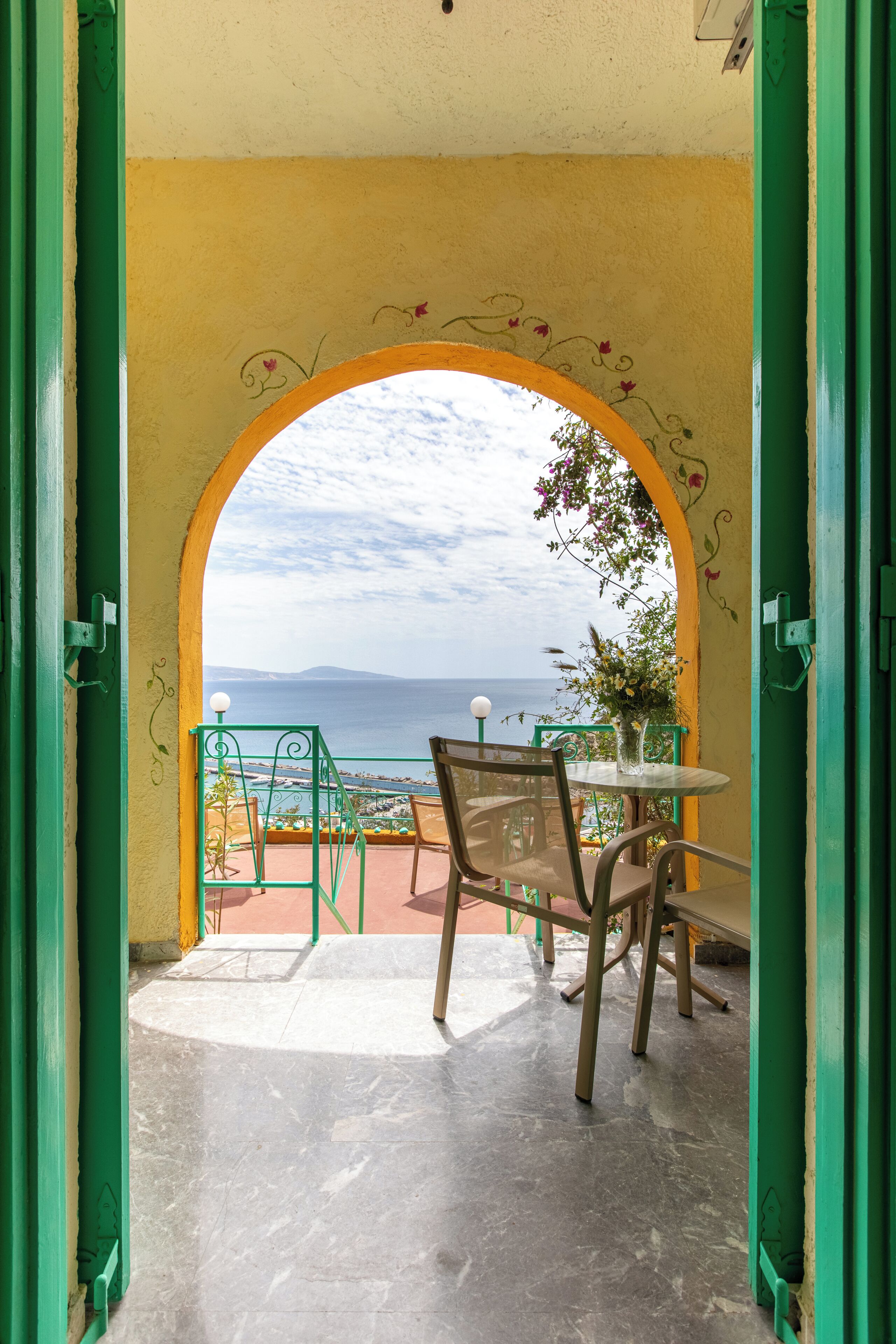 An open doorway leading to a terrace with a table and chairs, a view of the sea, and a yellow wall with floral decorations.