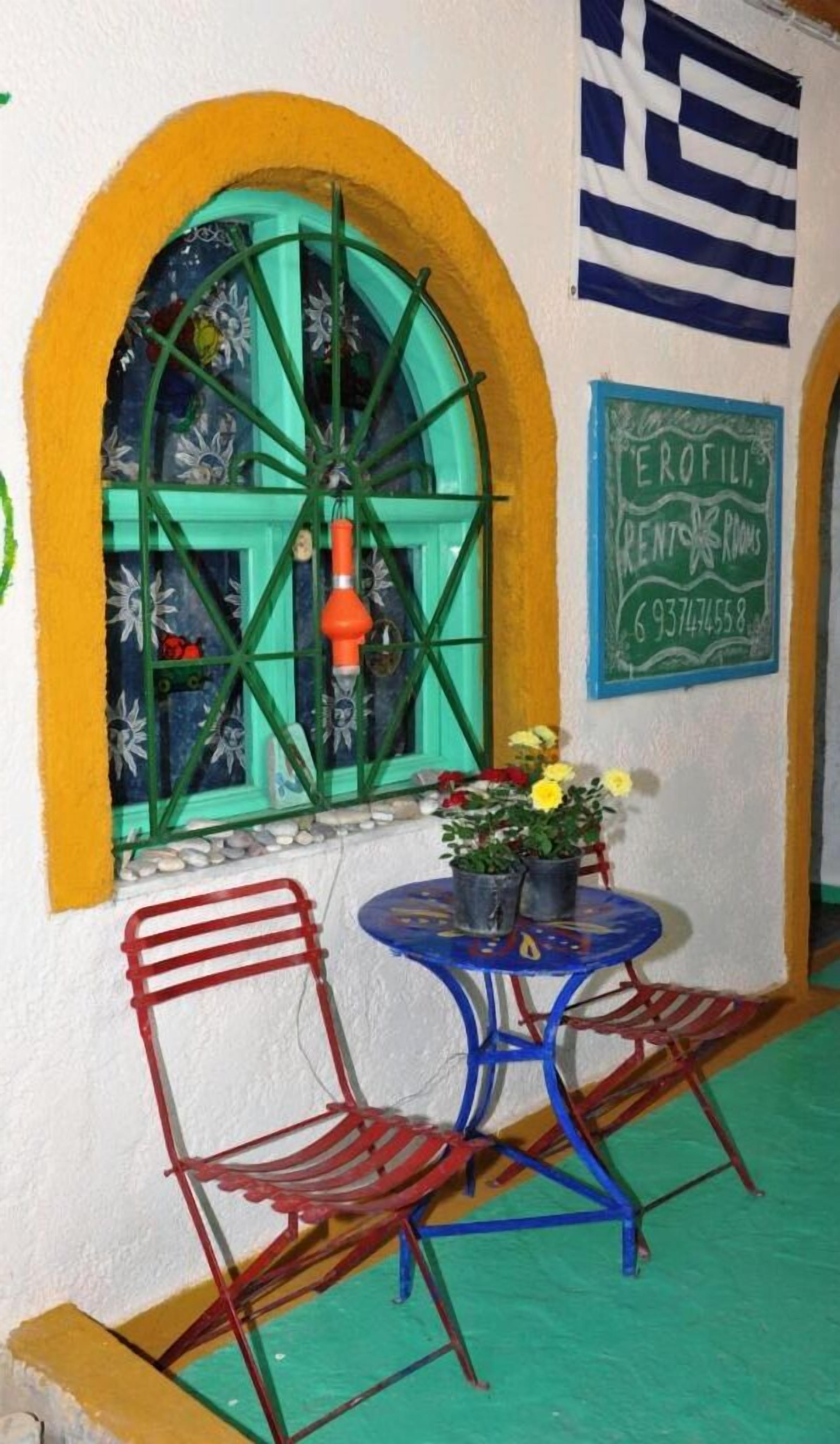 Un pequeño espacio al aire libre con dos sillas y una mesa, una ventana con marco verde y detalles decorativos, y un letrero que indica 'Erofile Rent-A-Rooms' con un número de teléfono.