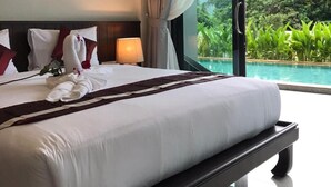 Una cama grande con ropa blanca y un cabecero oscuro, una mesita de noche con lámpara, y vistas a una piscina y vegetación.