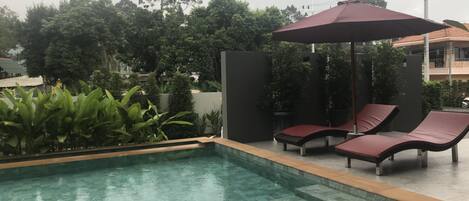 Una piscina rectangular con borde de madera, rodeada de sillas de descanso y un paraguas rojo.