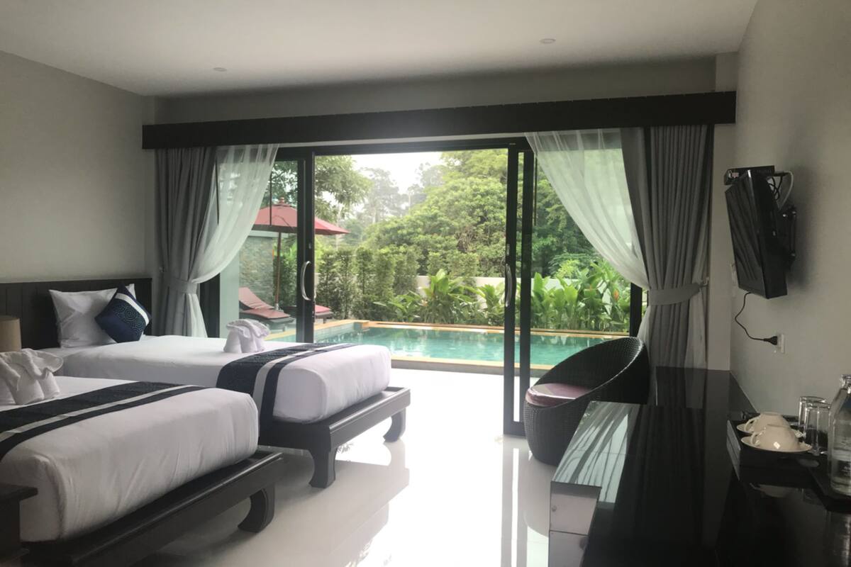 A hotel room with two beds, a TV, a dining table, and a view of a swimming pool and greenery.