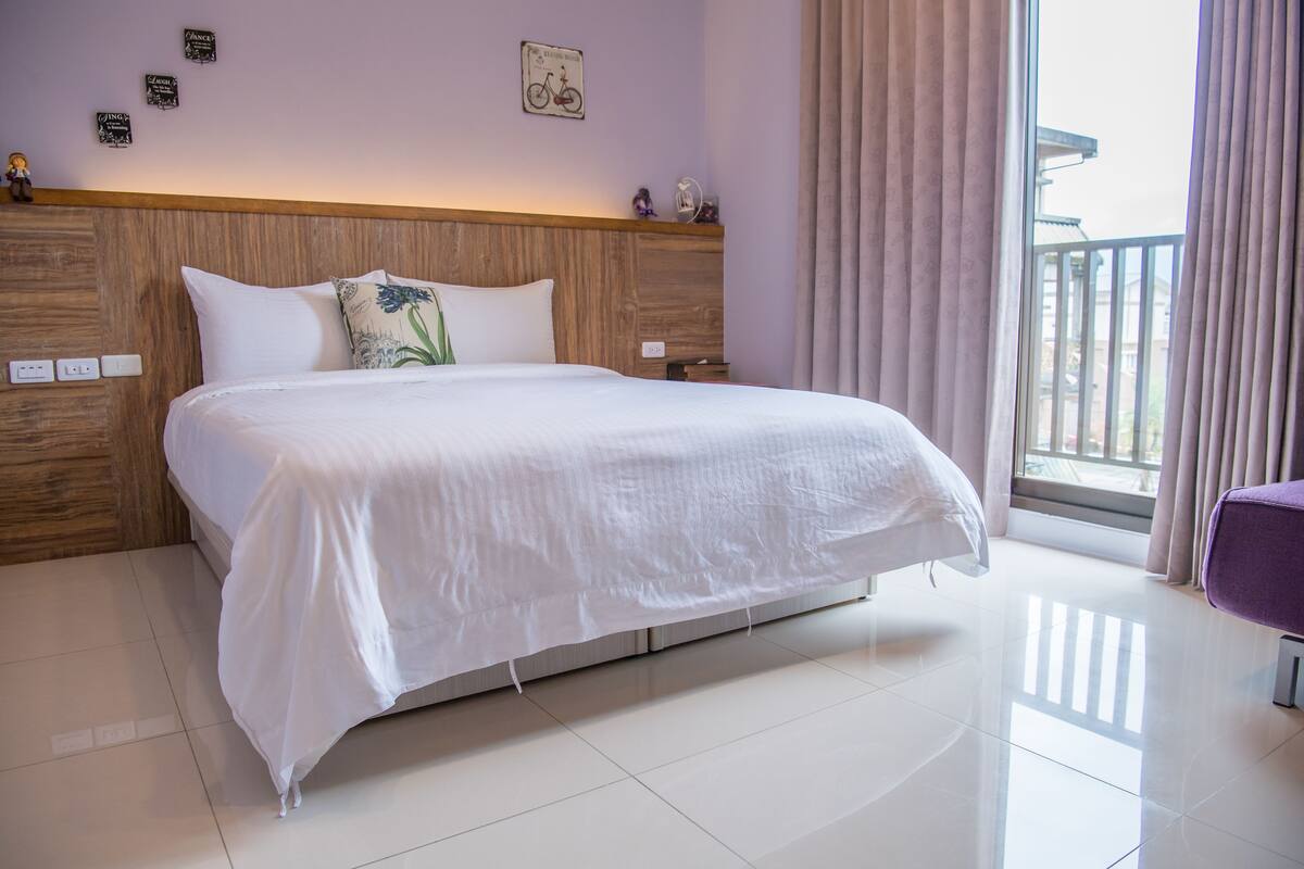 Uma cama bem arrumada com lençóis brancos, cabeceira de madeira e vista para uma varanda através de uma janela com cortinas.