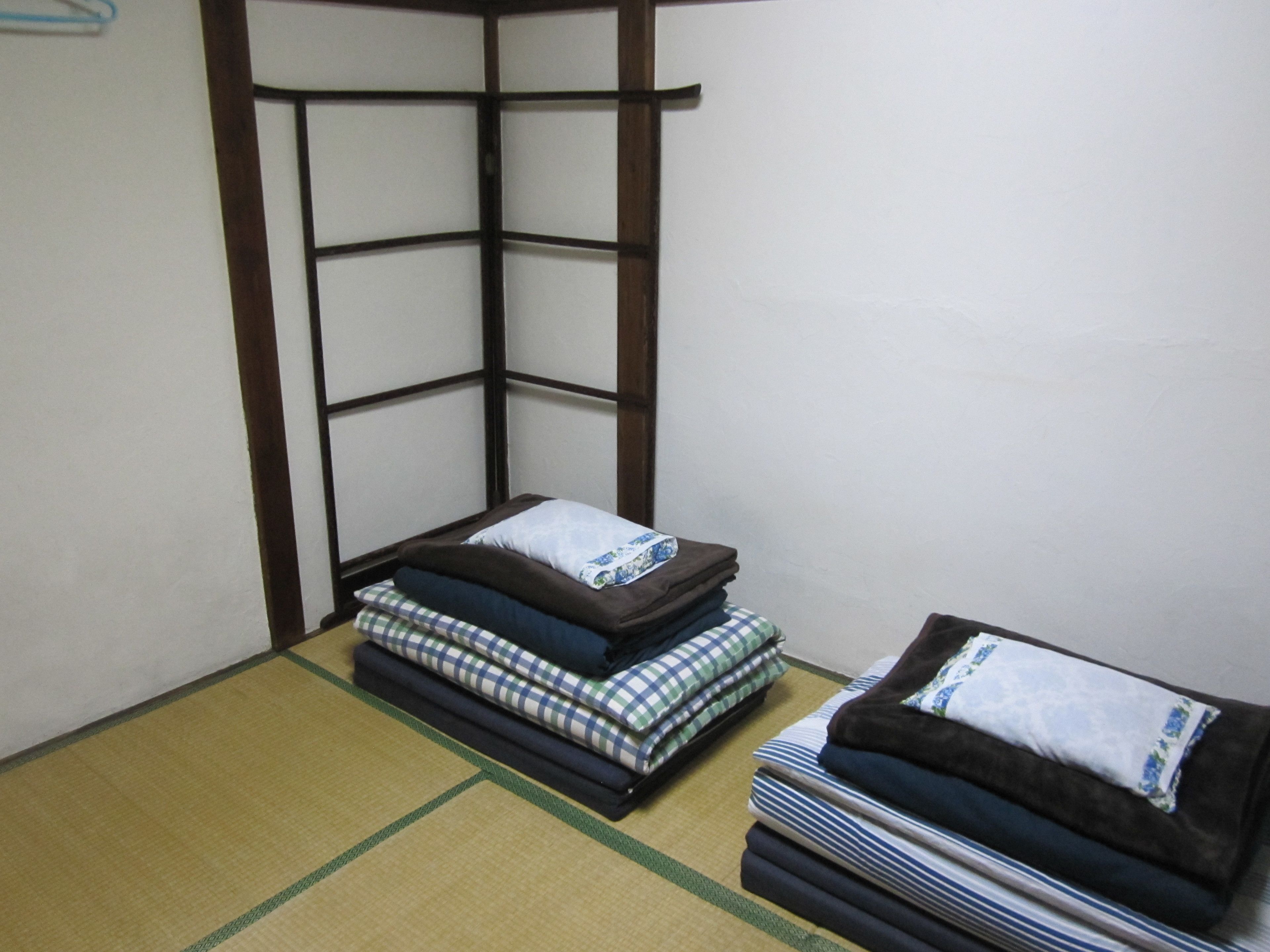 A corner room with tatami flooring, a low wooden bed frame, and neatly stacked bedding.